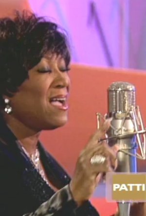 A wide shot of a busy recording session in a large studio, a young singer at a microphone with headphones, musicians and sound engineers visible behind glass, warm amber lighting, documentary style, thoughtful mood